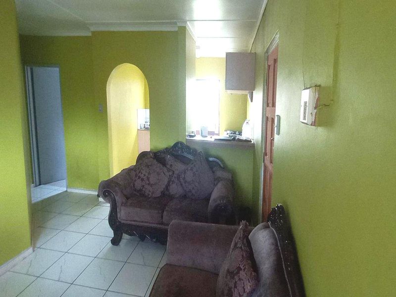 A living area featuring tiled flooring and an arched entryway leading to the kitchen, with dated wall finishes and visible electrical fixtures.