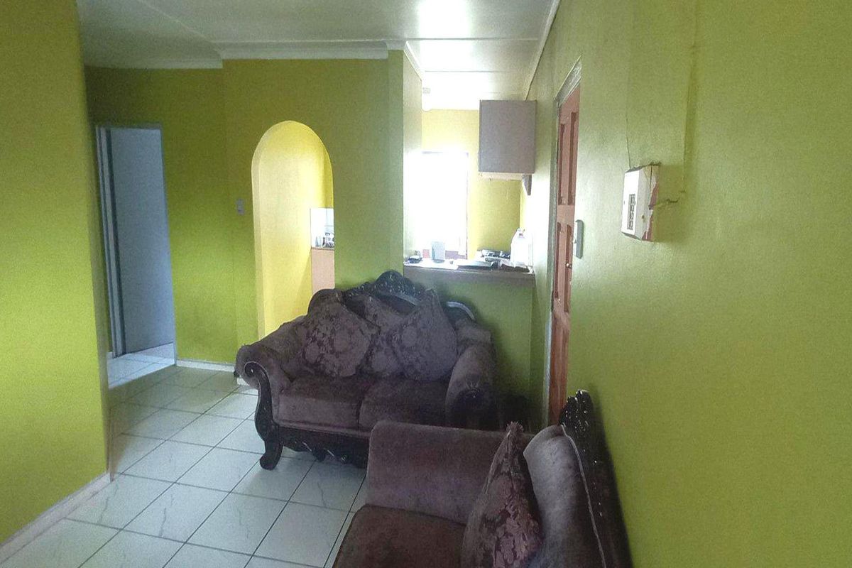 A living area featuring tiled flooring and an arched entryway leading to the kitchen, with dated wall finishes and visible electrical fixtures.