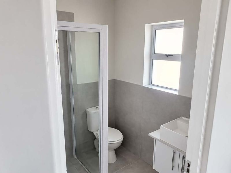 A clean, modern bathroom featuring a shower enclosure, a white vanity with basin, and neutral grey wall and floor tiling.