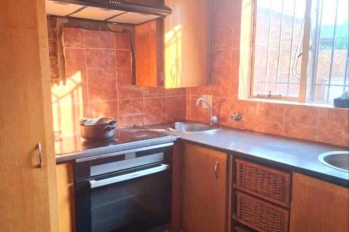 A compact kitchen featuring wooden cabinetry, an integrated oven with hob, extractor fan, and tiled backsplash with a window overlooking the exterior.