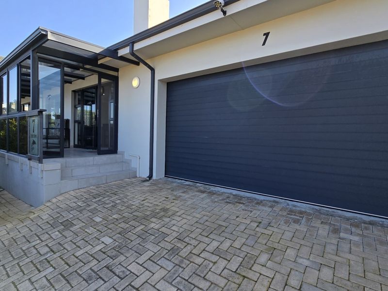 Front elevation featuring a modern dark garage door, paved driveway, and an enclosed glass-paneled patio entrance.