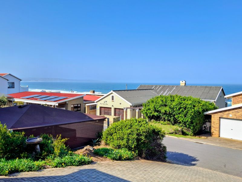 Elevated coastal view overlooking residential rooftops, paved driveways, and the ocean horizon under a clear blue sky.