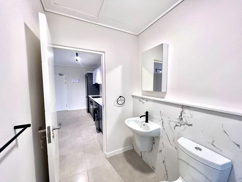 Modern bathroom featuring a wall-mounted basin with black fixtures, marble-look wall tiling, and a sleek mirror cabinet, leading into an open-plan kitchen area.