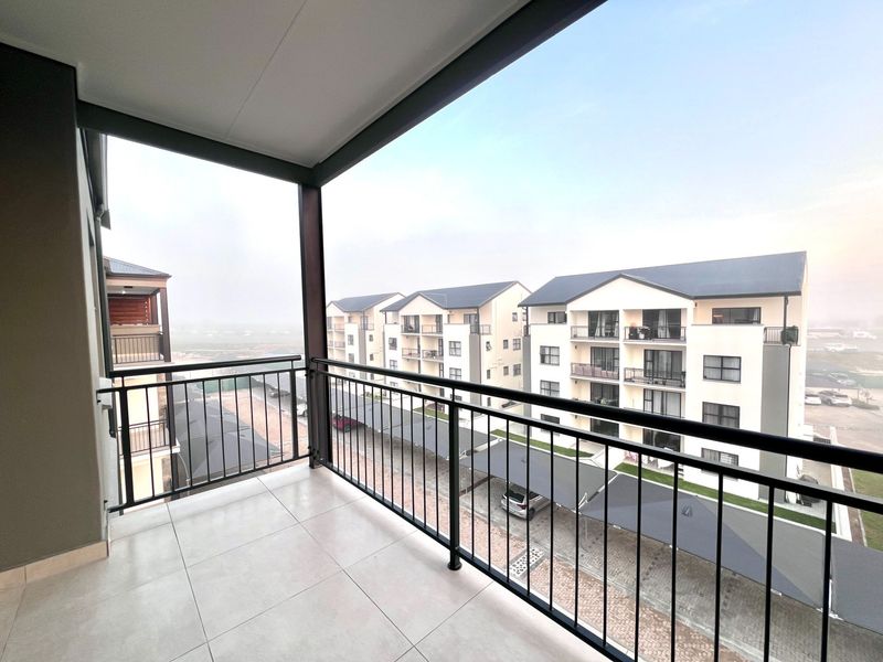 A clean, modern covered balcony with tiled flooring and a metal balustrade, offering an elevated view of the apartment complex and parking area.