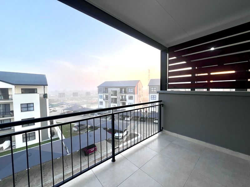 A clean, modern balcony featuring tiled flooring, a secure metal balustrade, and a privacy screen, offering an elevated view of the apartment complex.