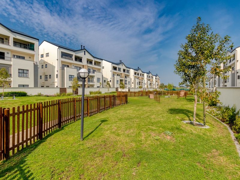 A well-maintained communal garden area featuring manicured lawns, perimeter fencing, and exterior lighting, set against a modern apartment complex.
