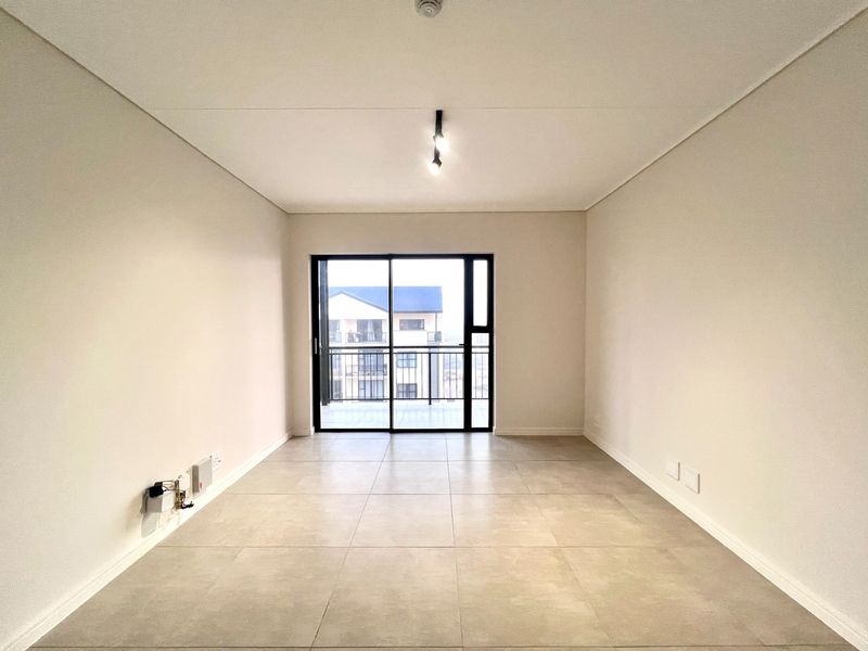 A bright, empty living area featuring neutral tiled flooring, modern ceiling lighting, and a sliding glass door providing access to an outdoor balcony.