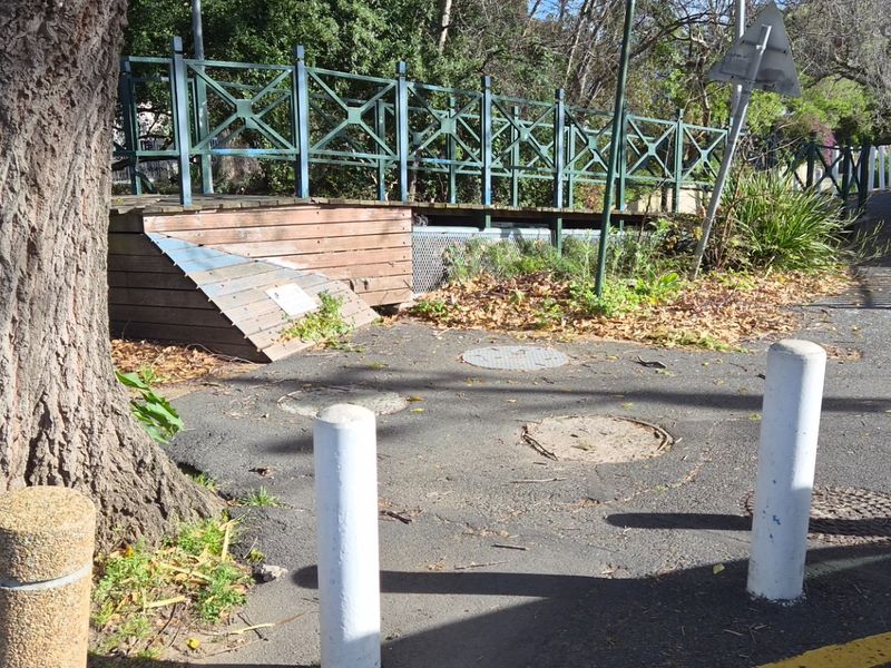 A street-level view showing a pedestrian bridge, asphalt pathway, and concrete bollards in a public outdoor area.