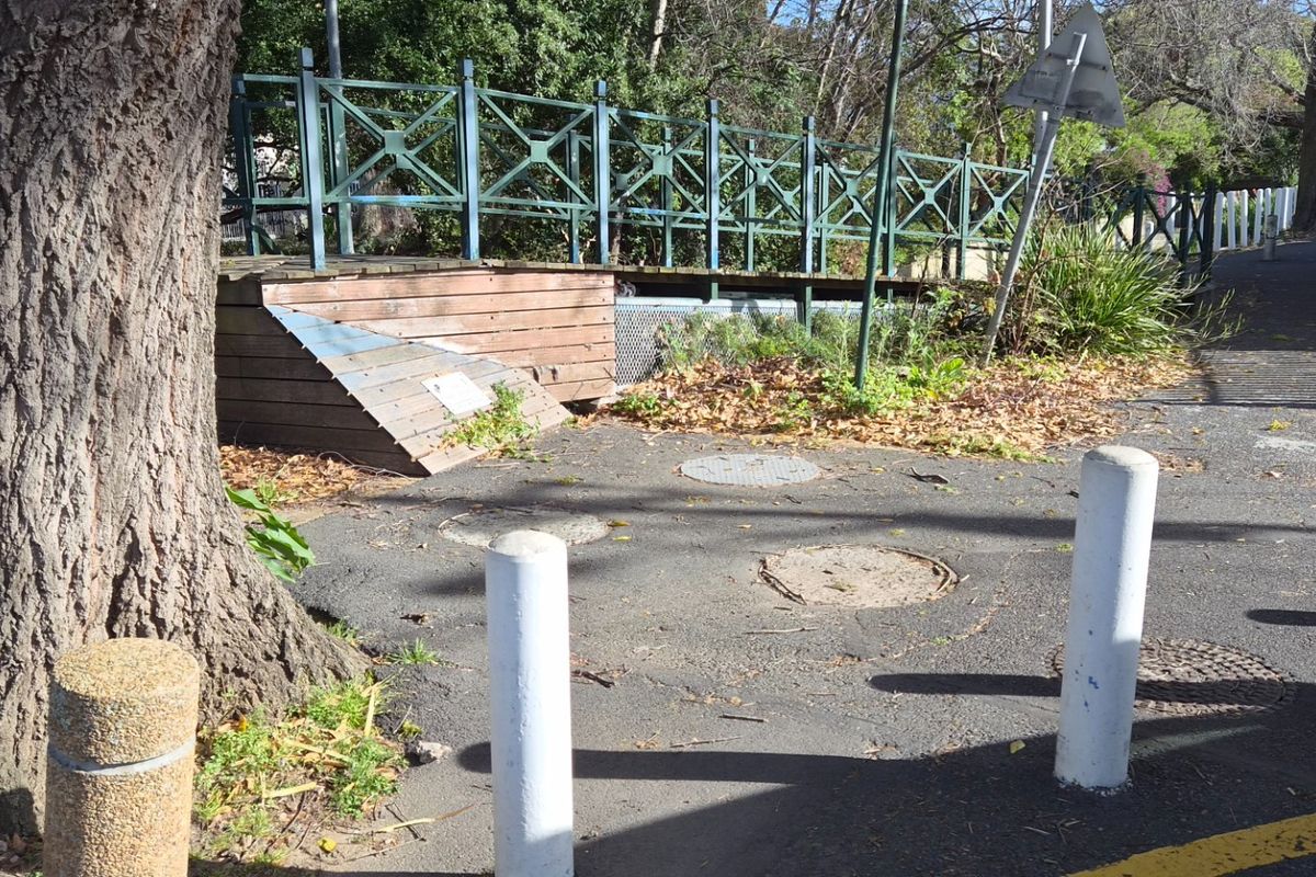 A street-level view showing a pedestrian bridge, asphalt pathway, and concrete bollards in a public outdoor area.