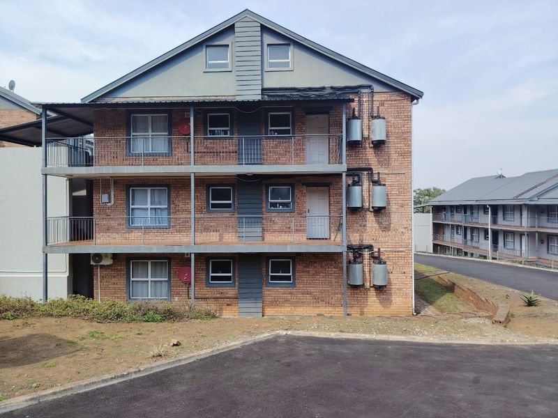 A three-story apartment building featuring face brick construction, external balcony access, and mounted geysers, situated within a paved residential complex.
