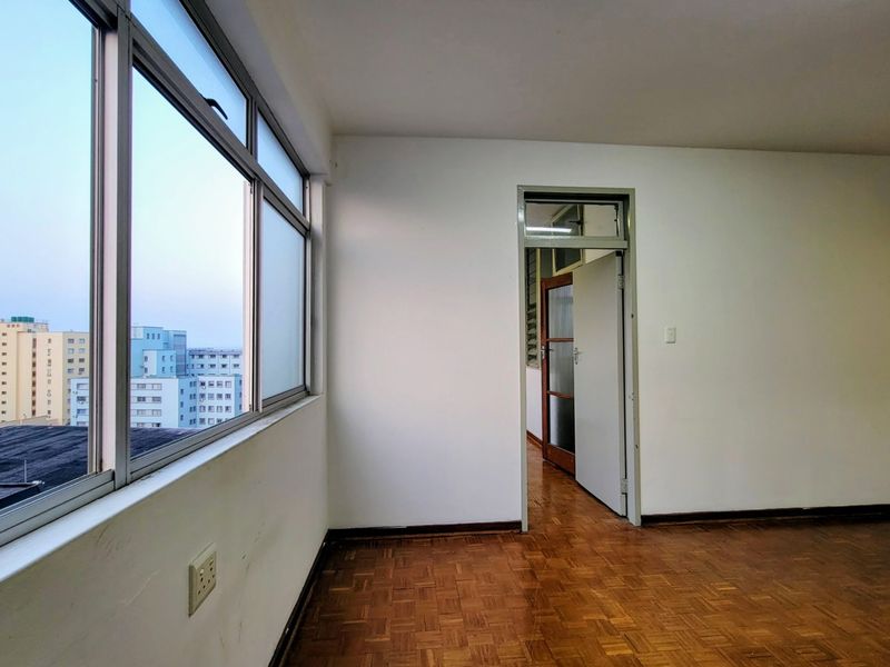 An empty living area featuring original parquet flooring, large aluminium-framed windows, and neutral walls, offering a blank canvas for interior design.