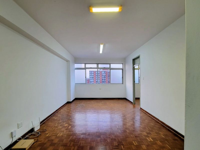 An empty living room featuring traditional parquet flooring, neutral walls, and large windows providing natural light.