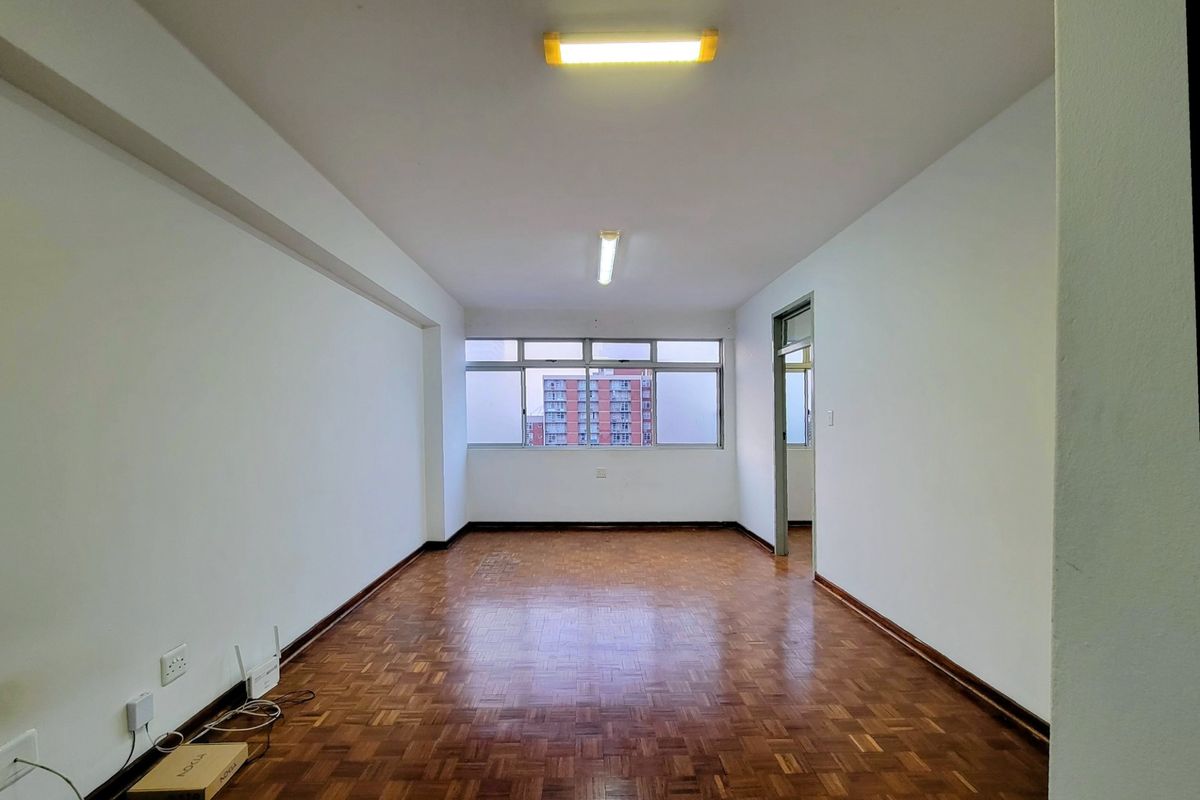 An empty living room featuring traditional parquet flooring, neutral walls, and large windows providing natural light.