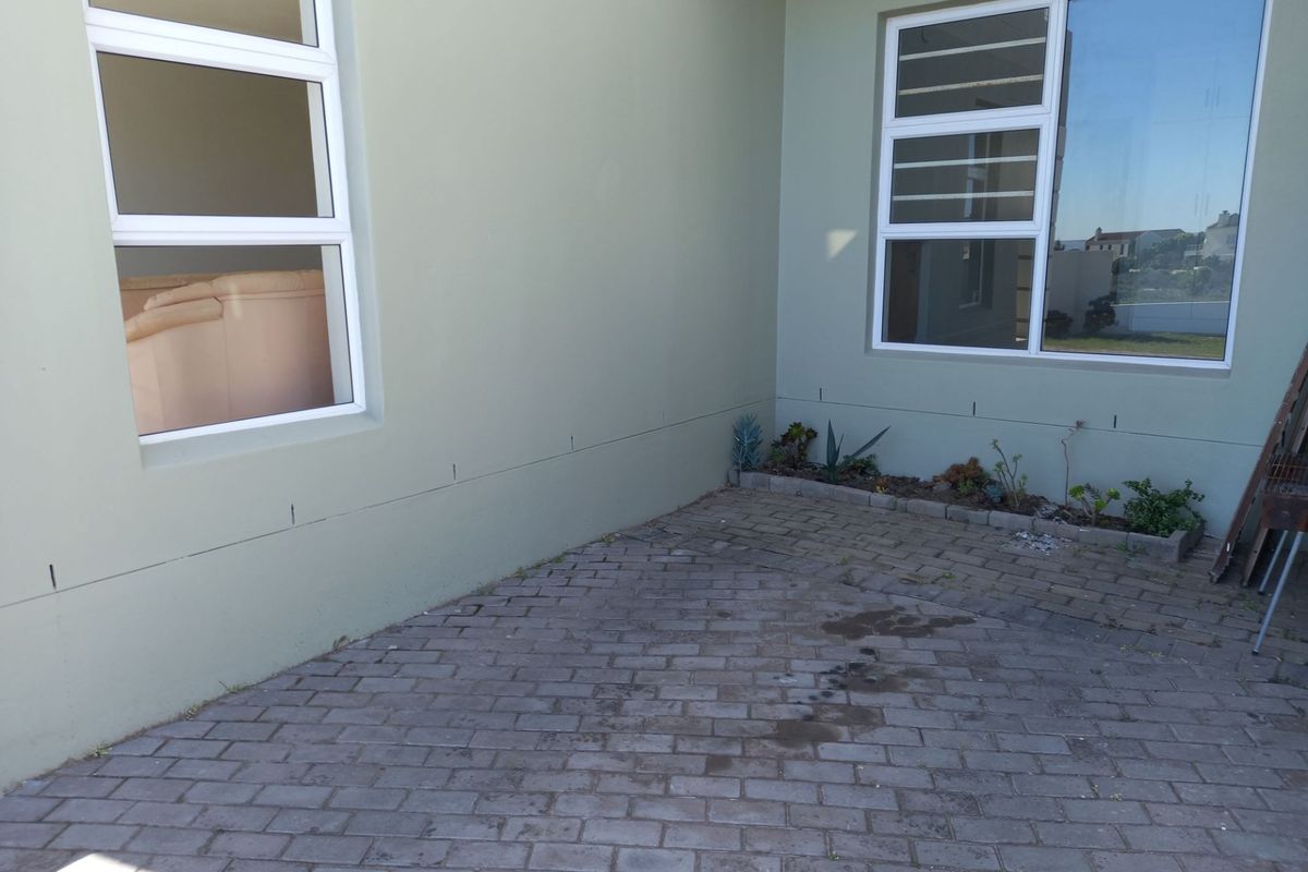 A paved rear patio area featuring light green exterior walls and aluminium window frames with security bars.