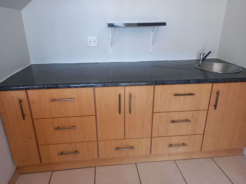 A functional scullery or utility area featuring wood-finish cabinetry, a dark stone countertop, a stainless steel sink, and tiled flooring.