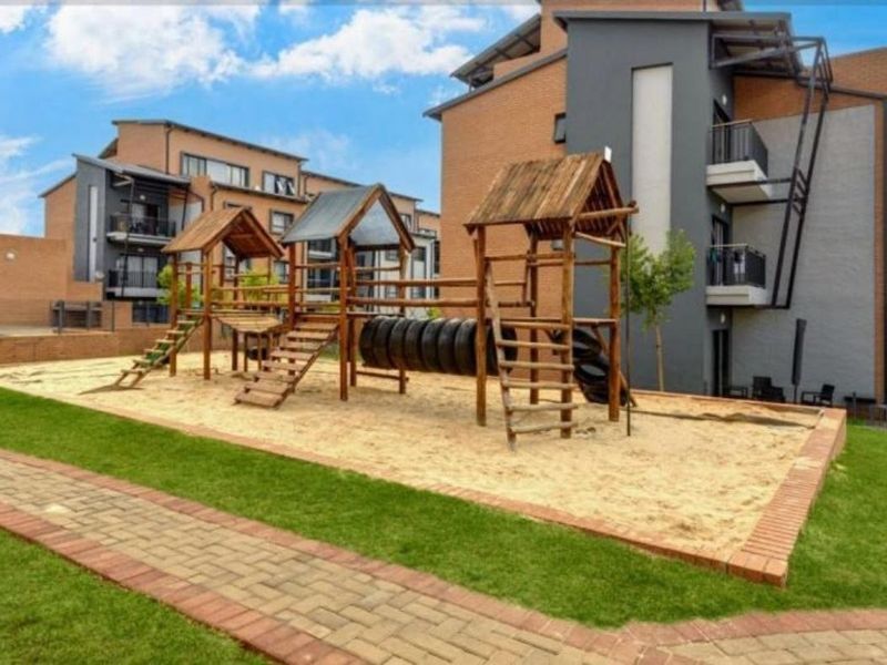 Communal outdoor area featuring a well-maintained children's playground with sand pit, set against a modern multi-story apartment complex with paved walkways.