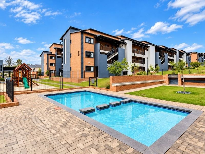 Communal outdoor area featuring a swimming pool with stepping stones, paved deck, landscaped lawn, and a children's playground within a secure apartment complex.