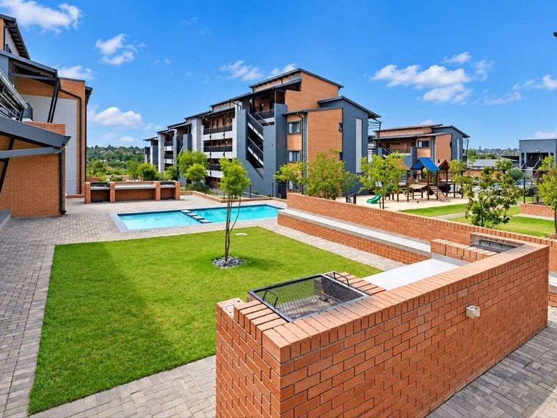 Communal outdoor area featuring a swimming pool, brick-built braai facilities, paved patio, and landscaped garden within a modern apartment complex.