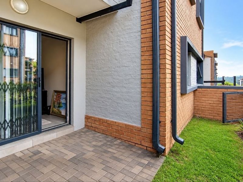 A neat ground-floor patio area featuring paved flooring, a sliding glass door with security gate, and direct access to a small garden space.