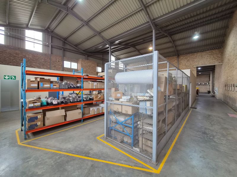 Spacious industrial warehouse area featuring exposed steel roof trusses, concrete flooring, secure metal mesh storage cage, and heavy-duty industrial shelving units.