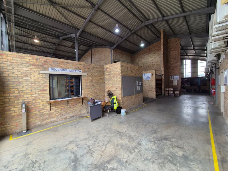 Industrial warehouse space featuring exposed brick walls, high steel roof trusses, concrete flooring, and a dedicated receiving and dispatch service window.