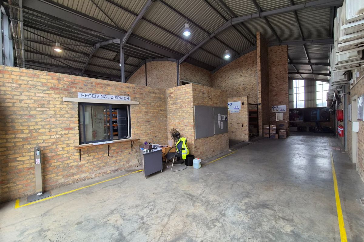 Industrial warehouse space featuring exposed brick walls, high steel roof trusses, concrete flooring, and a dedicated receiving and dispatch service window.