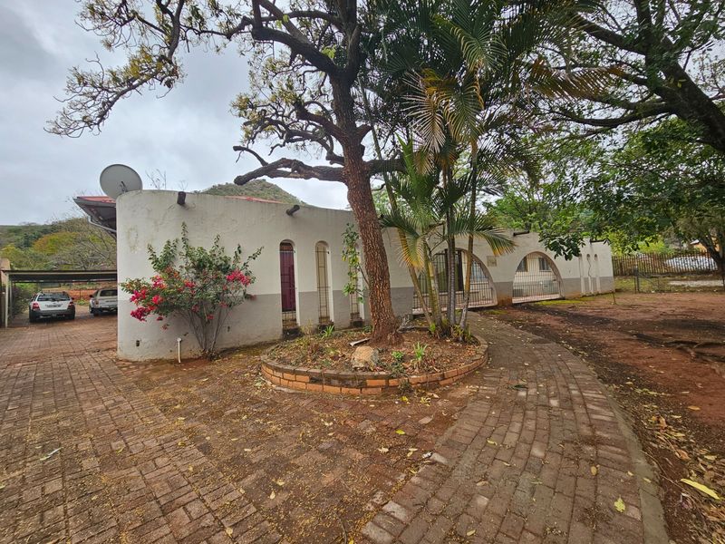 Front exterior view of the property featuring a paved driveway, carport, and arched architectural details with mature trees and landscaping.