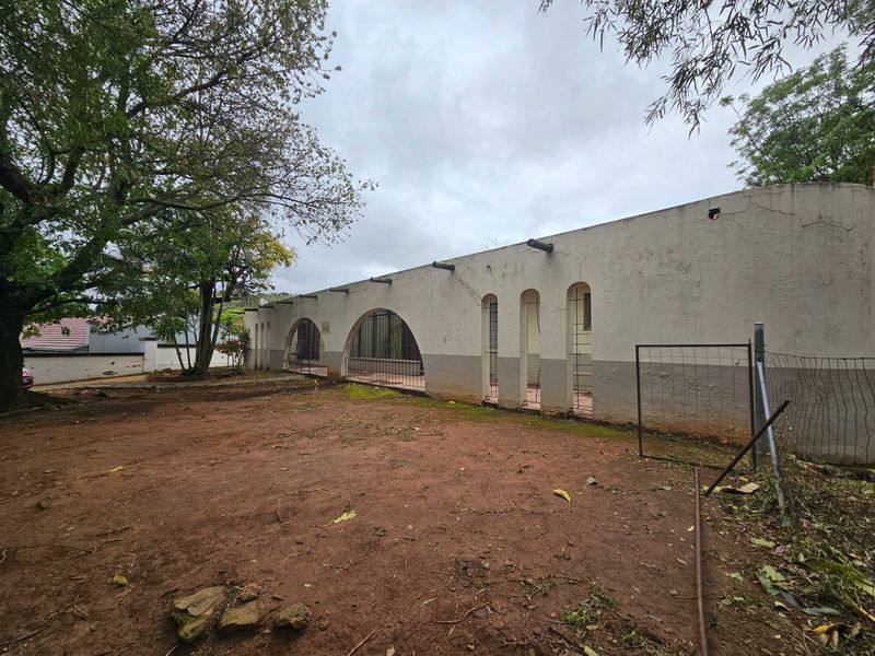 Rear exterior view of the property featuring arched architectural openings, exposed timber beams, and a large unlandscaped yard area requiring maintenance.