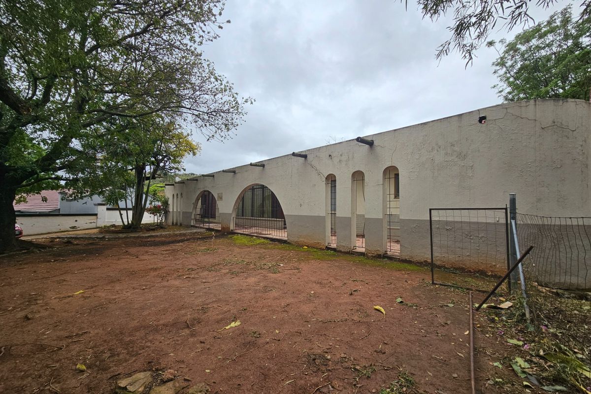 Rear exterior view of the property featuring arched architectural openings, exposed timber beams, and a large unlandscaped yard area requiring maintenance.