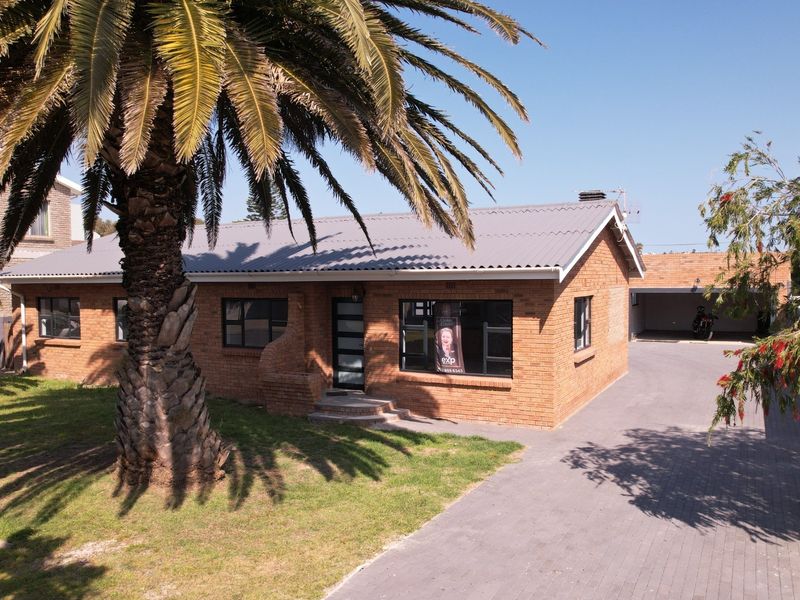 A well-maintained single-story brick house featuring a corrugated metal roof, a paved driveway, and a detached garage, set on a landscaped residential lot.