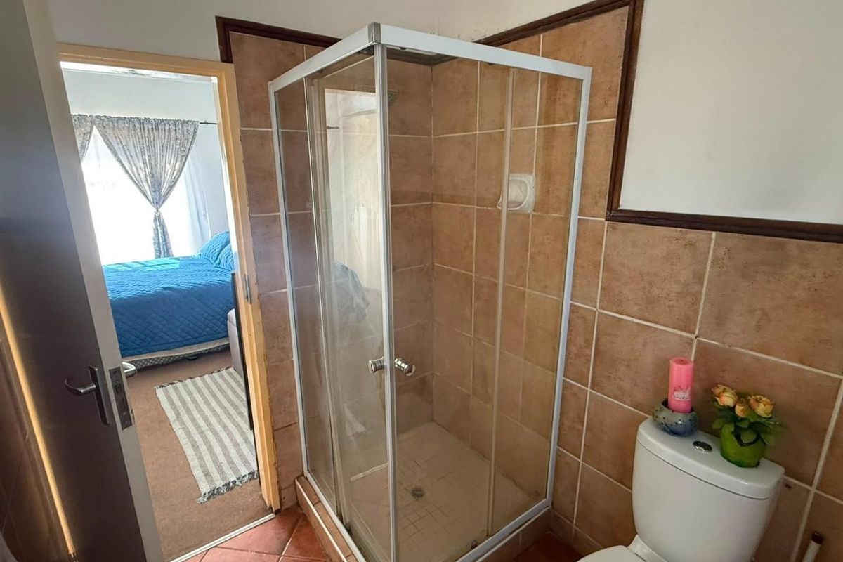 A compact bathroom featuring a corner shower cubicle, ceramic wall and floor tiling, and a standard toilet, with a view into the adjacent bedroom.