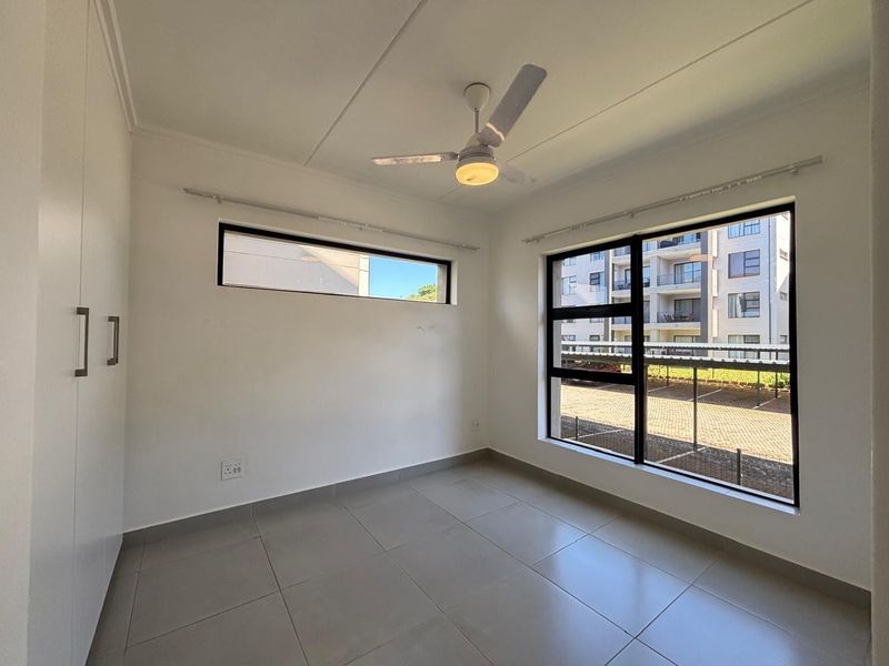 A bright, empty bedroom featuring neutral tiled flooring, built-in cupboards, a ceiling fan, and large windows providing natural light.