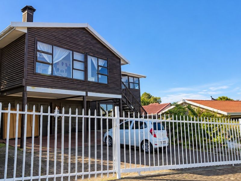 Front view of a two-story timber-clad house featuring an elevated deck, external staircase, and a secure gated driveway entrance.