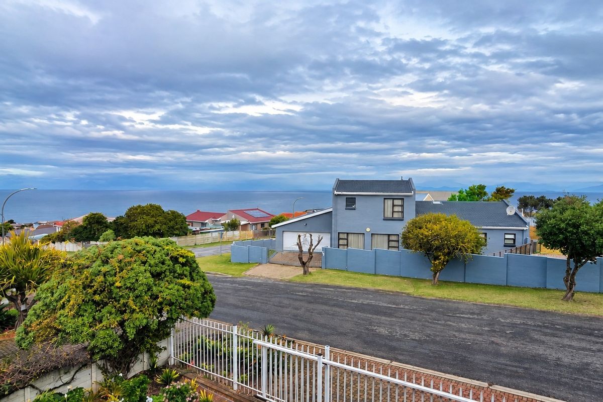 An elevated aerial view of a residential property featuring a blue exterior, perimeter wall, and driveway, set against a scenic coastal backdrop.