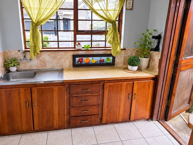 A functional scullery area featuring wooden cabinetry, a stainless steel sink, tiled splashback, and direct access to an outdoor space.