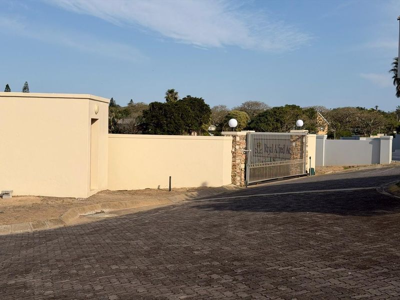 A secure entrance to the residential estate featuring a sliding gate, stone-clad pillars, and perimeter walling along a paved access road.