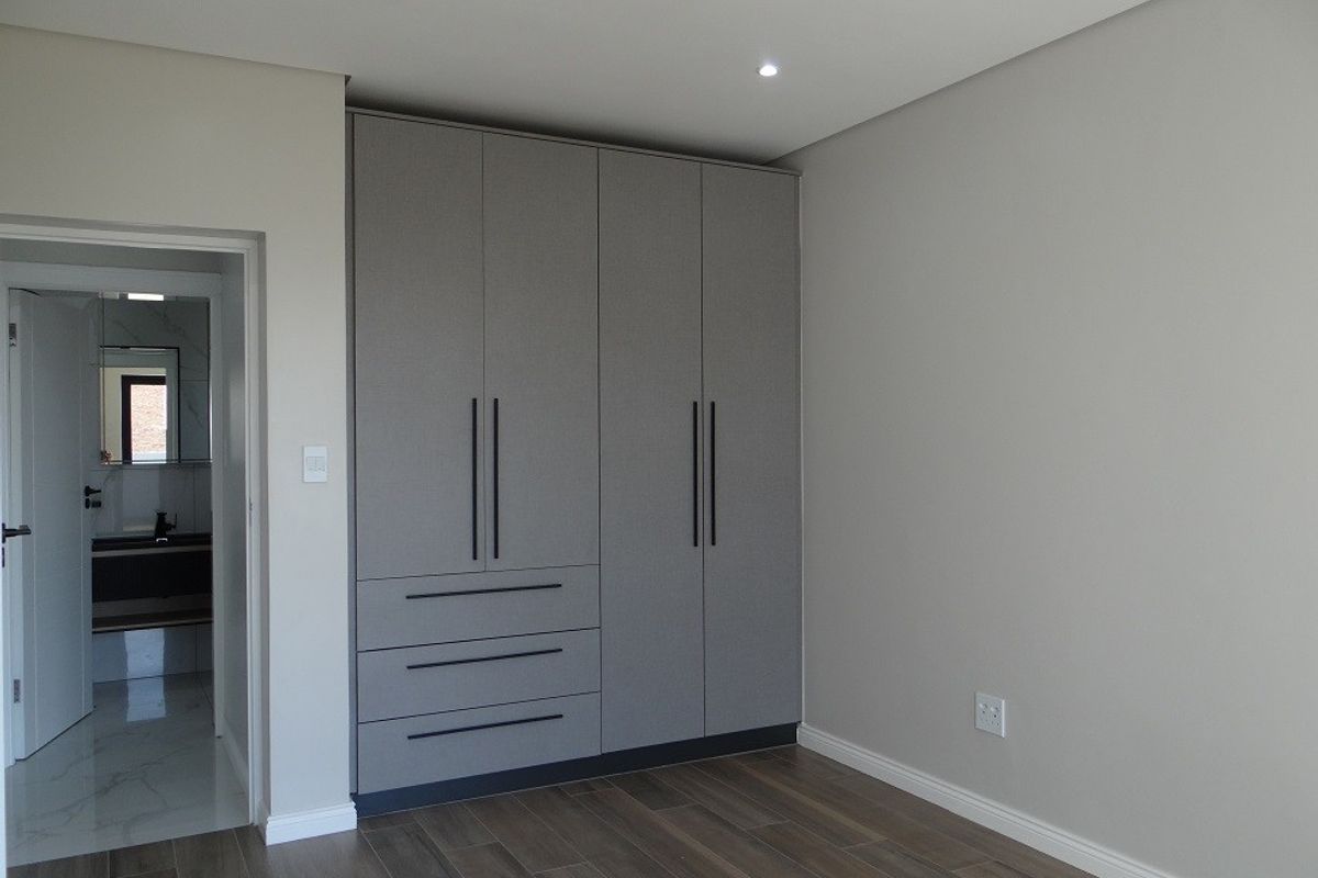A modern, empty bedroom featuring neutral grey built-in cupboards, recessed downlights, and wood-look laminate flooring, with an adjacent view into a tiled bathroom.
