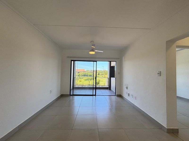 An empty, well-lit living room featuring neutral tiled flooring, a ceiling fan, and sliding glass doors leading to an outdoor balcony.