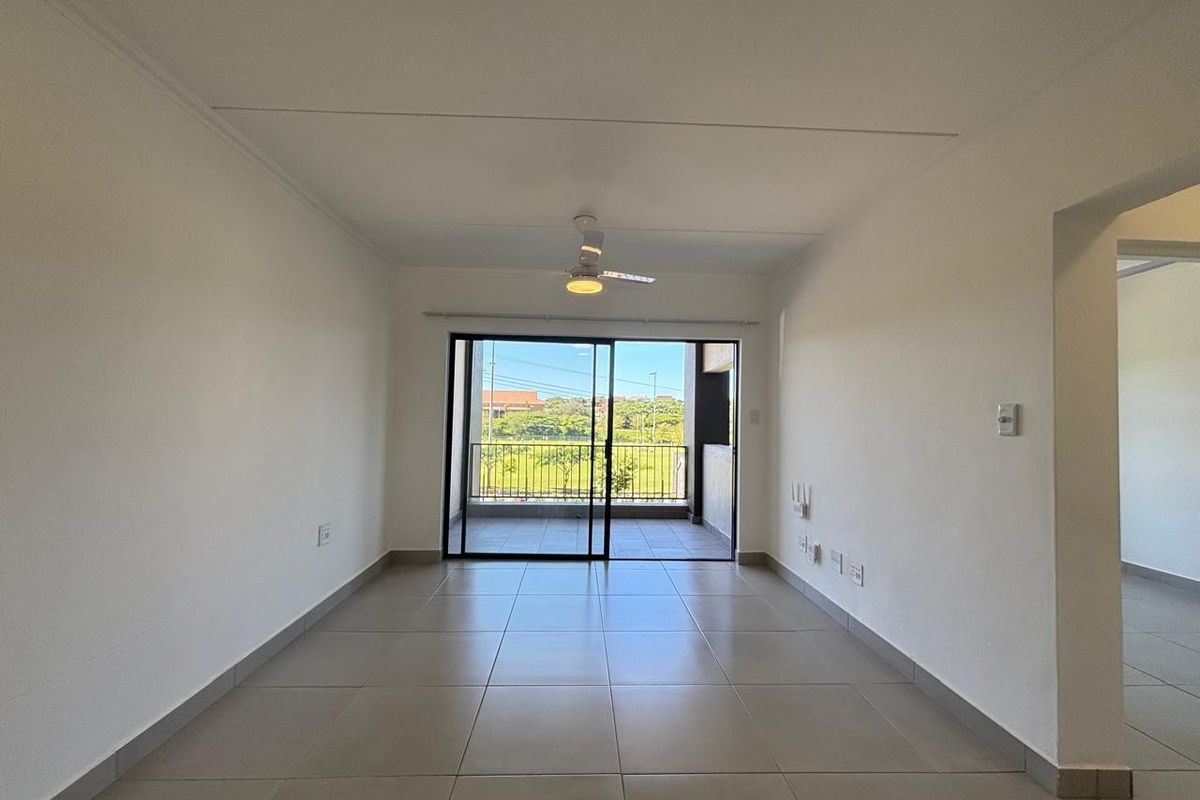An empty, well-lit living room featuring neutral tiled flooring, a ceiling fan, and sliding glass doors leading to an outdoor balcony.