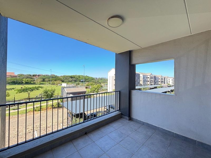 A spacious covered balcony featuring tiled flooring and a metal balustrade, offering pleasant views of the surrounding estate and green space.