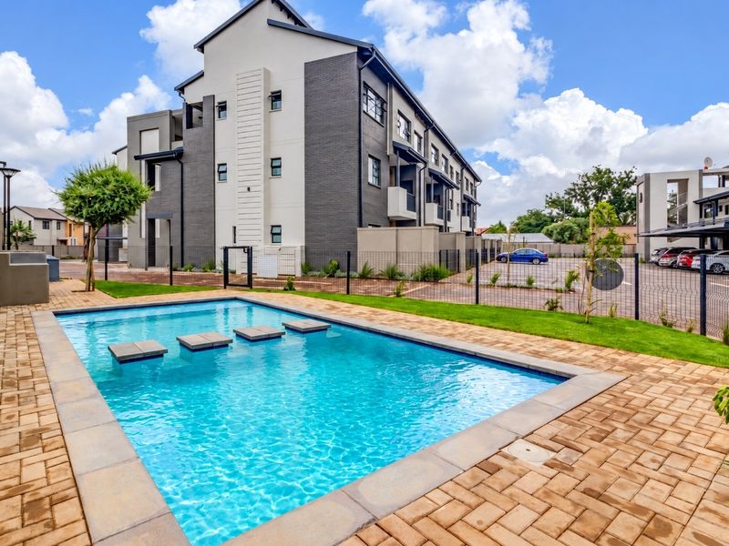 A well-maintained communal swimming pool area featuring stepping stones, paved decking, and landscaped surroundings within a modern apartment complex.