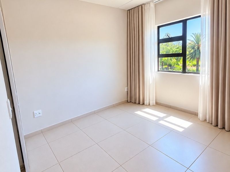 A bright, empty bedroom featuring neutral tiled flooring, a built-in cupboard, and large windows with curtains, offering a clean and well-maintained living space.