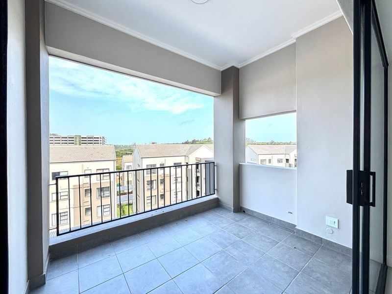 A clean, covered balcony featuring tiled flooring and a metal balustrade, offering an open view of the surrounding apartment complex.