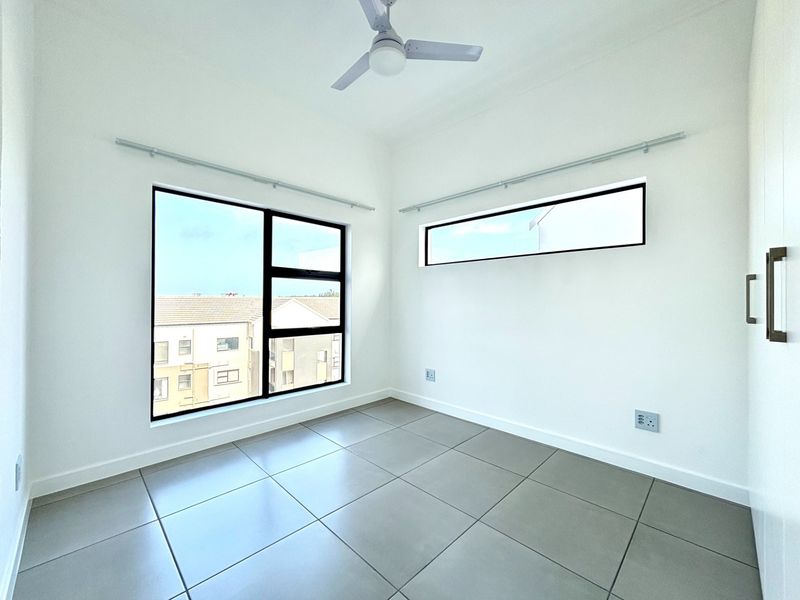 A bright, modern bedroom featuring neutral tiled flooring, built-in cupboards, a ceiling fan, and large windows with aluminium frames.
