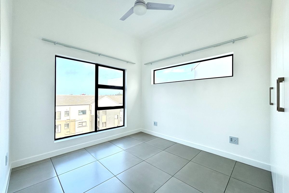 A bright, modern bedroom featuring neutral tiled flooring, built-in cupboards, a ceiling fan, and large windows with aluminium frames.