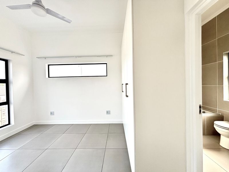 A bright, modern bedroom featuring neutral tiled flooring, built-in cupboards, a ceiling fan, and an en-suite bathroom.