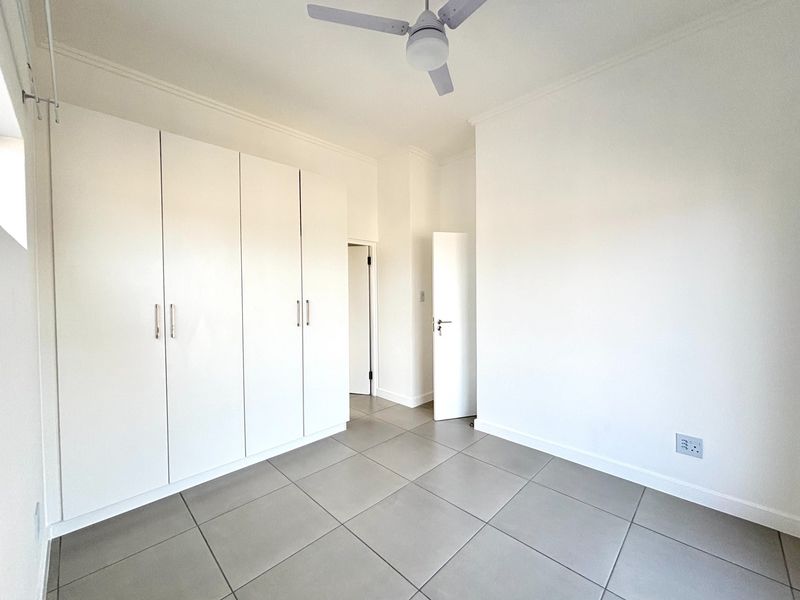 A bright, empty bedroom featuring neutral tiled flooring, built-in white cupboards, and a ceiling fan, offering a clean and versatile living space.