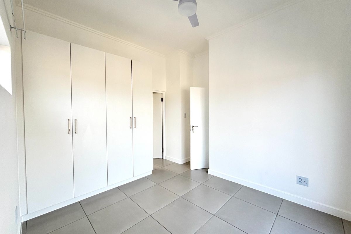 A bright, empty bedroom featuring neutral tiled flooring, built-in white cupboards, and a ceiling fan, offering a clean and versatile living space.