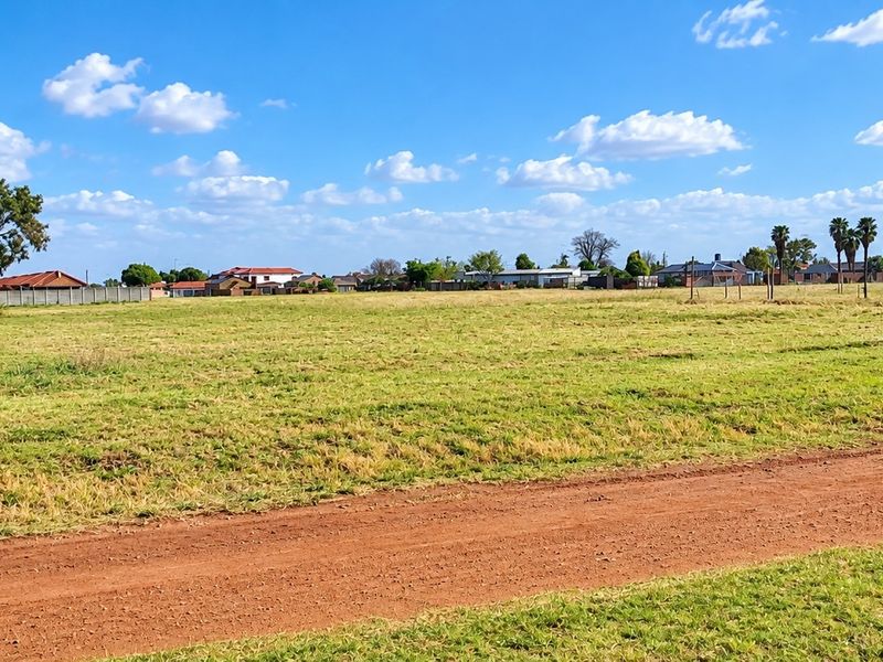 A wide-angle view of a vacant residential plot featuring a dirt road access and perimeter fencing, set within a developed suburban neighborhood.