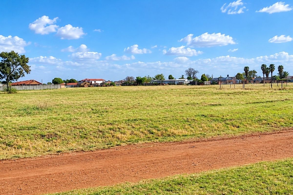 A wide-angle view of a vacant residential plot featuring a dirt road access and perimeter fencing, set within a developed suburban neighborhood.